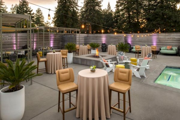 An outdoor patio setup with high tables, barstools, lounge chairs, string lights, a fire pit, and potted plants next to a small pool, surrounded by trees.