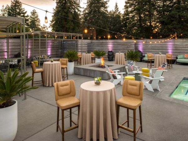 An outdoor patio setup with high tables, barstools, lounge chairs, string lights, a fire pit, and potted plants next to a small pool, surrounded by trees.