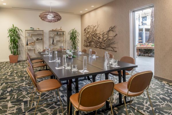 A modern conference room with a long table, chairs, plants, and wall decor; there is an open door leading outside in the background.