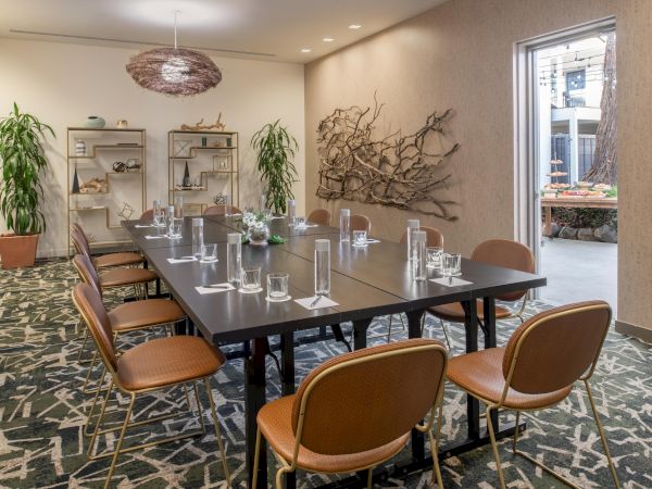 A modern conference room with a long table, chairs, plants, and wall decor; there is an open door leading outside in the background.