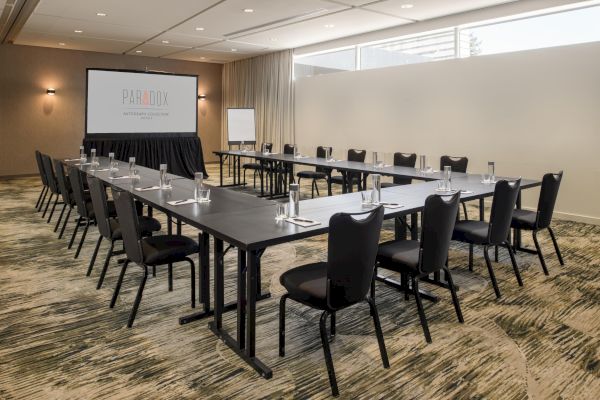 A conference room set up with U-shaped tables, chairs, a projector screen displaying 'Paradox', and a flip chart.