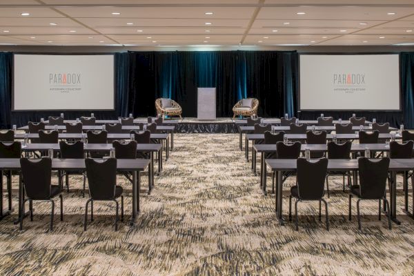 This image shows a conference room set up with rows of tables and chairs facing a stage with two chairs and two screens displaying 