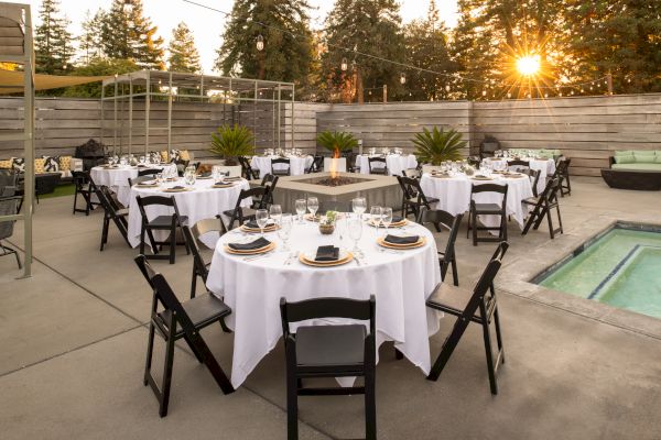 An outdoor event setup with multiple round tables covered in white tablecloths, surrounded by black chairs, situated near a jacuzzi and string lights.