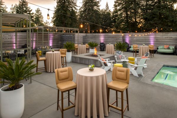 This image shows an outdoor patio setup with round tables, bar stools, string lights, potted plants, and a small pool, surrounded by a wooden fence.