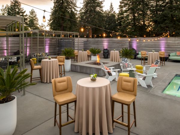 This image shows an outdoor patio setup with round tables, bar stools, string lights, potted plants, and a small pool, surrounded by a wooden fence.