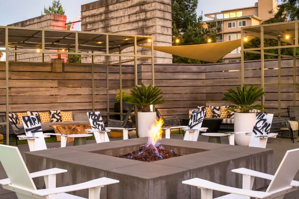 An outdoor seating area with a central fire pit, white chairs, potted plants, and wooden decor. It looks like a cozy social space.