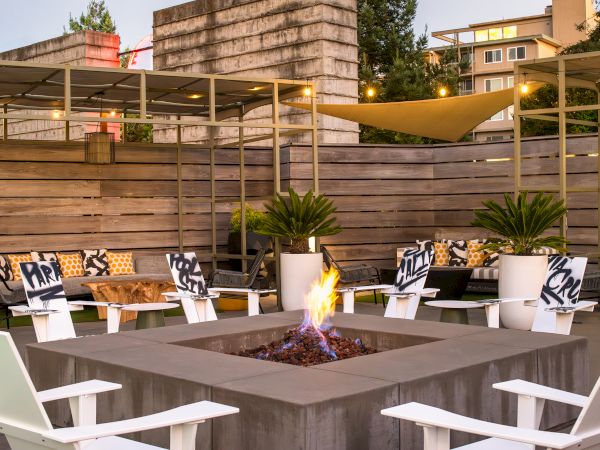 An outdoor seating area with a central fire pit, white chairs, potted plants, and wooden decor. It looks like a cozy social space.