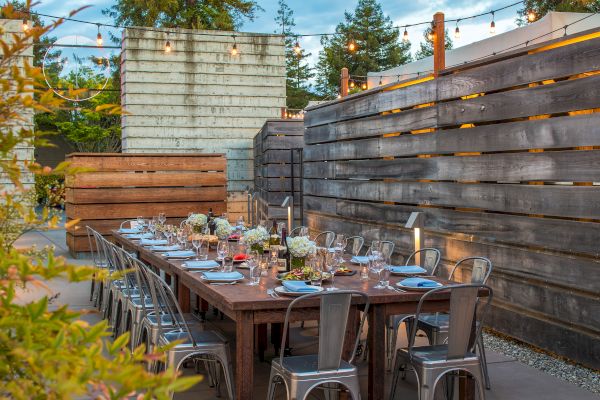 An outdoor dining area with a long table set with flowers, glasses, and plates, surrounded by metal chairs, under string lights, with wooden walls.