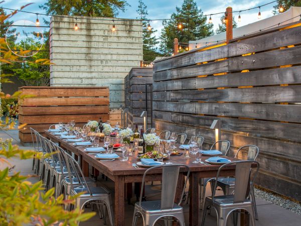 An outdoor dining area with a long table set with flowers, glasses, and plates, surrounded by metal chairs, under string lights, with wooden walls.