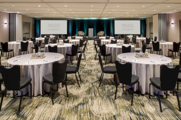 A conference room with round tables, black chairs, and notepads. There are two screens displaying 