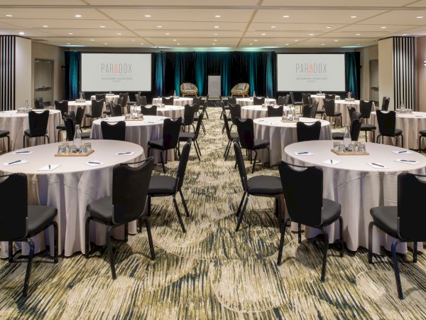 A conference room with round tables, black chairs, and notepads. There are two screens displaying "PARADOX" on either side of the stage.