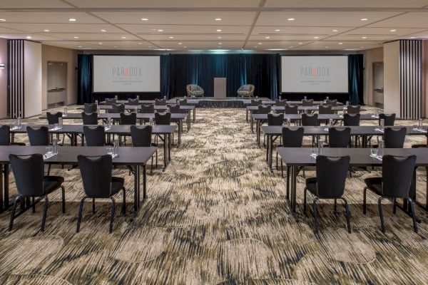 A conference room with multiple rows of tables and chairs, two large screens, and a podium in front. The room is set for a presentation.