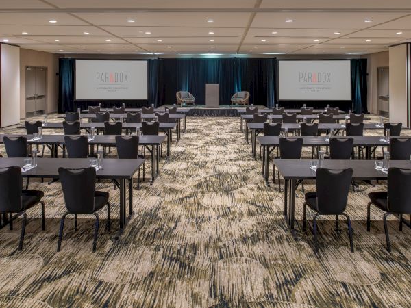 A conference room with multiple rows of tables and chairs, two large screens, and a podium in front. The room is set for a presentation.