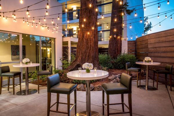 The image shows an outdoor patio area with tall tables, chairs, string lights, and floral centerpieces, creating a cozy and inviting ambiance.