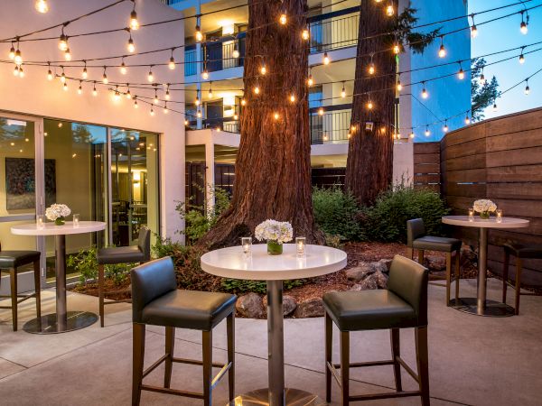 The image shows an outdoor patio area with tall tables, chairs, string lights, and floral centerpieces, creating a cozy and inviting ambiance.