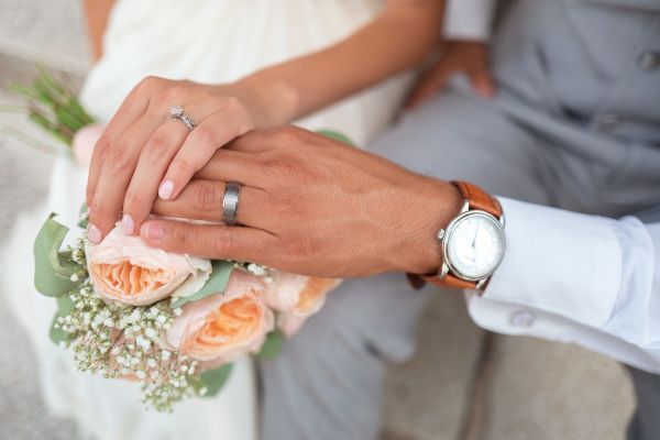 A close-up image of two people holding hands, one wearing a wedding ring, with a bouquet of flowers nearby and a watch on one wrist.