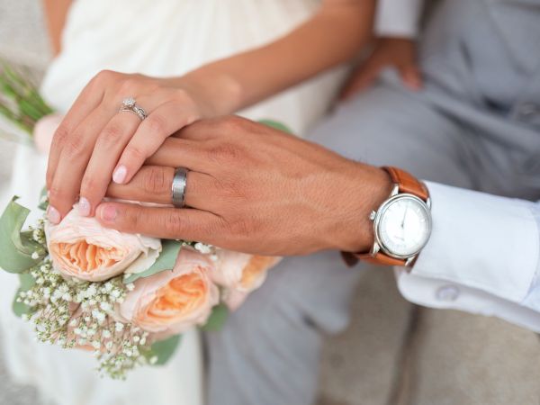 A close-up image of two people holding hands, one wearing a wedding ring, with a bouquet of flowers nearby and a watch on one wrist.