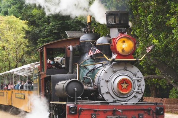 An old steam locomotive with passengers is chugging along a track, emitting steam, and passing through a green, wooded area.