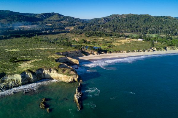 A scenic coastal landscape with rocky cliffs, sandy beach, and clear blue water, set against a backdrop of rolling hills and lush greenery.