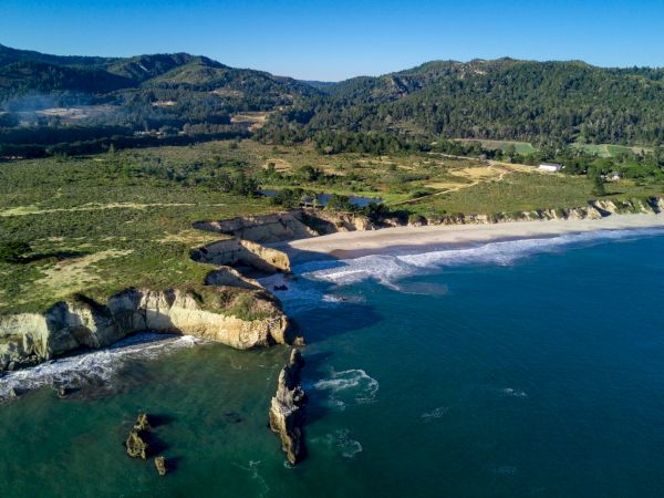 A scenic coastal landscape with rocky cliffs, sandy beach, and clear blue water, set against a backdrop of rolling hills and lush greenery.