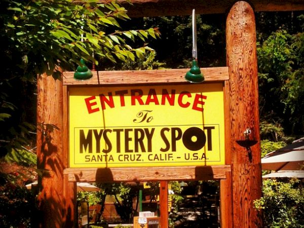 The image shows a sign that reads "Entrance to Mystery Spot, Santa Cruz, Calif. - U.S.A." hanging between rustic wooden posts.