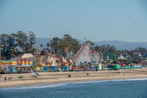 A lively beach scene with an amusement park featuring a variety of rides, including a roller coaster, and colorful buildings in the background.