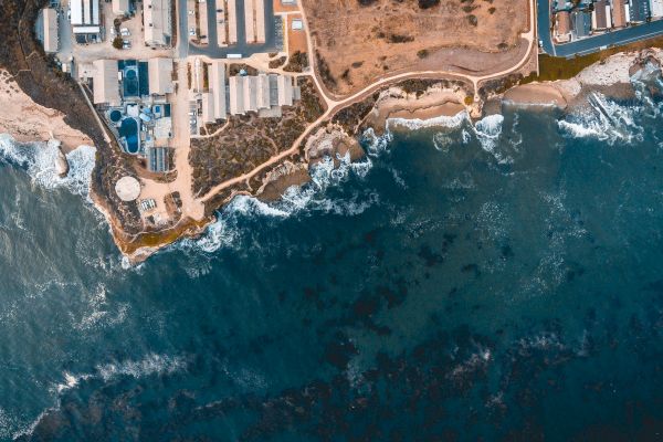 An aerial view of a coastal area featuring buildings, roads, and rocky cliffs meeting the ocean with waves crashing against the shore.