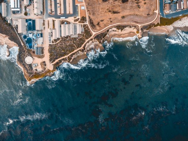 An aerial view of a coastal area featuring buildings, roads, and rocky cliffs meeting the ocean with waves crashing against the shore.