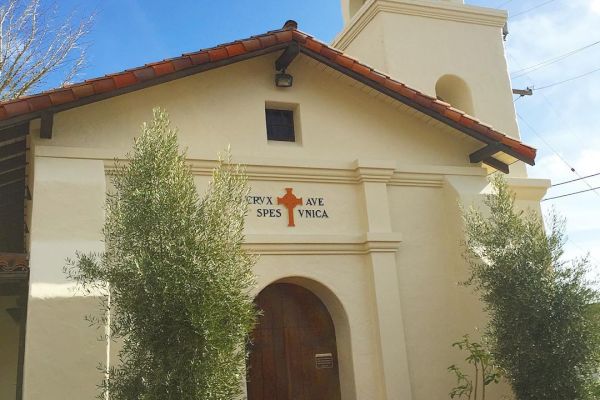 The image shows a small church with a beige facade, an arched wooden door, a bell tower, and a cross on top. The words 