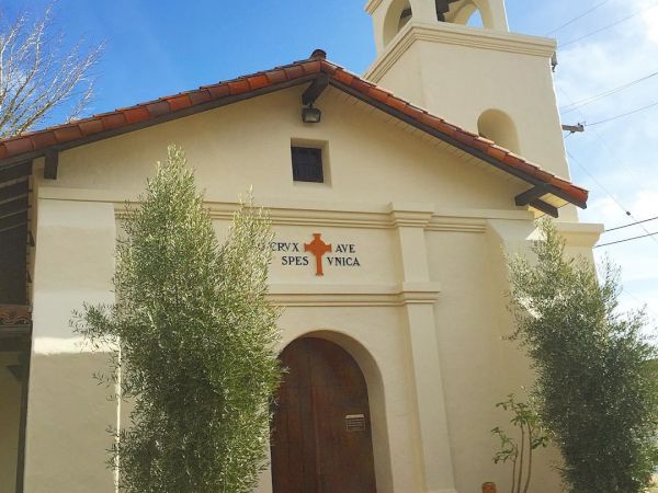The image shows a small church with a beige facade, an arched wooden door, a bell tower, and a cross on top. The words "Ora et Labora" are above the door.