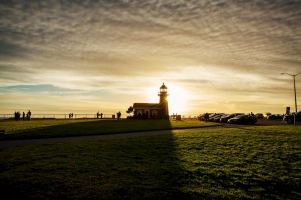 The image shows a small lighthouse at sunset, casting long shadows on the grass, with people and cars in the distance near the ocean.