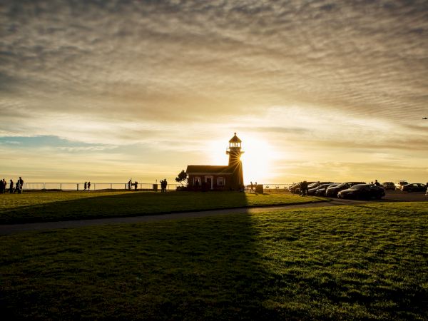 The image shows a small lighthouse at sunset, casting long shadows on the grass, with people and cars in the distance near the ocean.