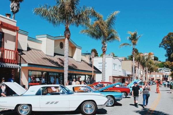 A street scene with classic cars parked along the side, people walking, and palm trees framing the buildings under a clear blue sky.