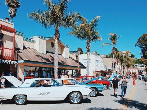 A street scene with classic cars parked along the side, people walking, and palm trees framing the buildings under a clear blue sky.