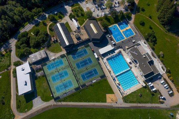 Aerial view of a sports complex with tennis courts, basketball courts, and swimming pools, surrounded by buildings and parking spaces.