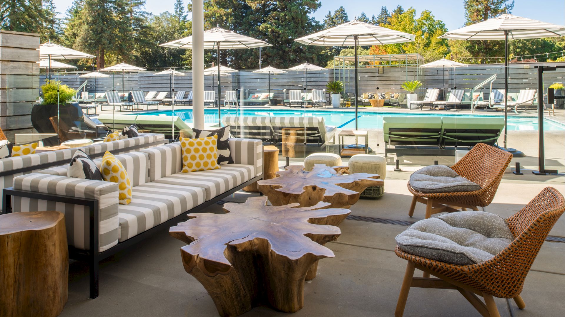 The image shows a stylish outdoor seating area with wooden tables, modern chairs, and a pool with umbrellas and lounge chairs in the background.