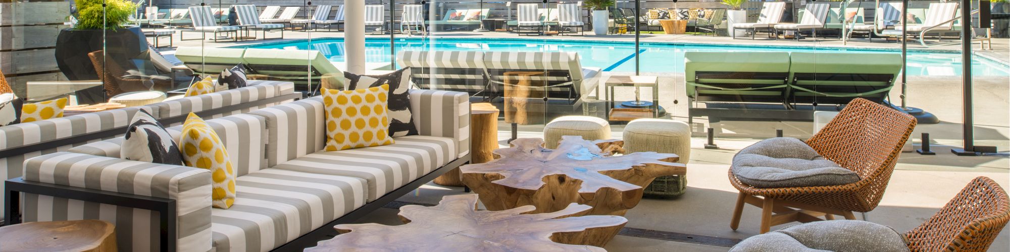 The image shows a stylish outdoor seating area with wooden tables, modern chairs, and a pool with umbrellas and lounge chairs in the background.