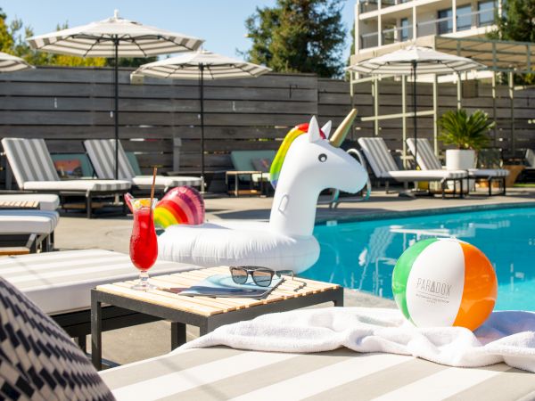 Poolside scene with lounge chairs, umbrellas, a unicorn float, a cocktail, sunglasses, a towel, and a beach ball by the water's edge.