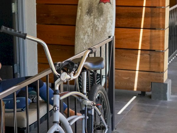 This image shows a bicycle parked next to a surfboard leaning against a wooden partition on a tiled patio.