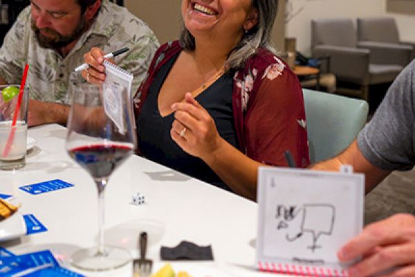 Three people are seated around a table, smiling and drawing on small notepads. There are drinks and snacks on the table.