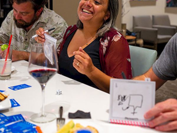 Three people are seated around a table, smiling and drawing on small notepads. There are drinks and snacks on the table.