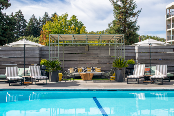 This image shows a pool area with lounge chairs, umbrellas, plants, and a seating area under a pergola, surrounded by a wooden fence and trees.