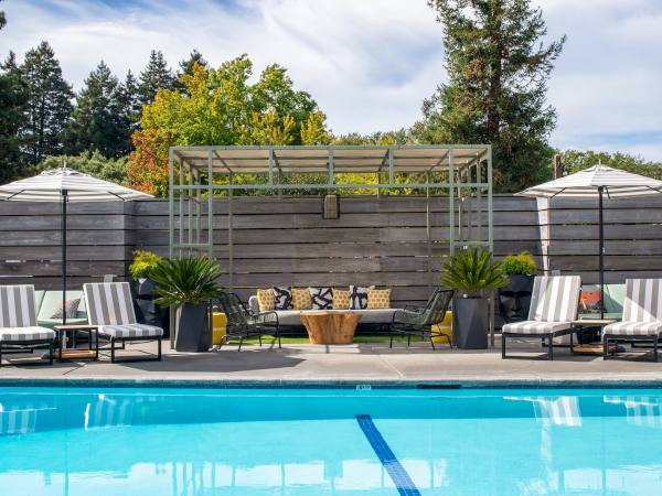 This image shows a pool area with lounge chairs, umbrellas, plants, and a seating area under a pergola, surrounded by a wooden fence and trees.
