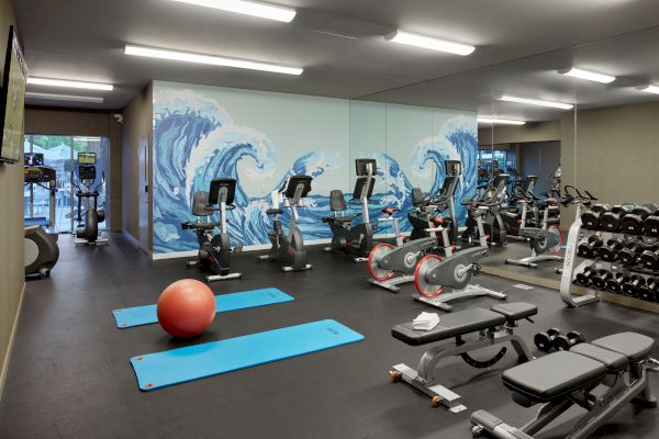 A gym room with exercise equipment including stationary bikes, treadmills, dumbbells, benches, yoga mats, and a large mural of ocean waves.