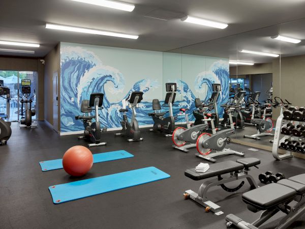 A gym room with exercise equipment including stationary bikes, treadmills, dumbbells, benches, yoga mats, and a large mural of ocean waves.