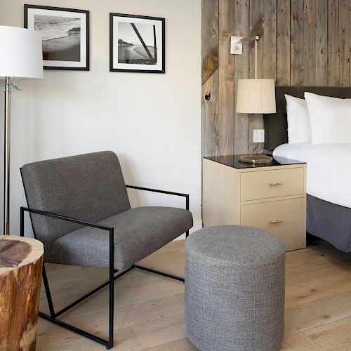 This image shows a cozy bedroom with a bed, a bedside table, a floor lamp, an armchair, a wooden side table, and two framed photos on the wall.