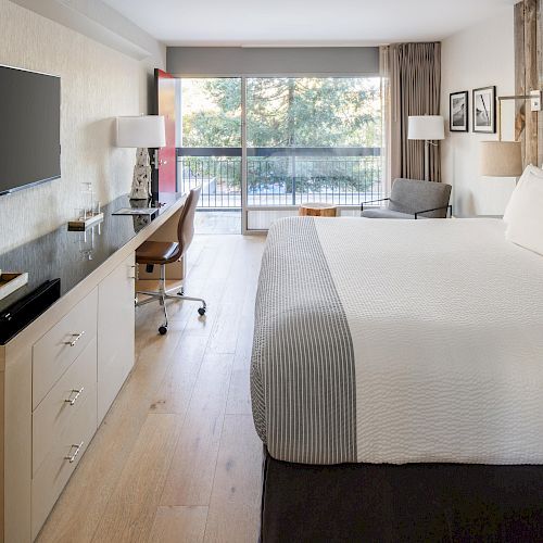 The image shows a modern hotel room with a large bed, flat-screen TV, desk, chair, and a window with a view of trees outside.