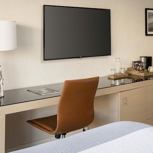 A modern room with a desk featuring a lamp, magazine, brown chair, TV, two pictures, and a beverage tray with a coffee maker and cups.