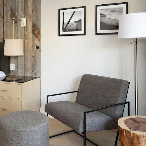 A cozy room with a bed, nightstand, lamp, gray chair, ottoman, wooden stump table, floor lamp, and two framed black-and-white photos on the wall.