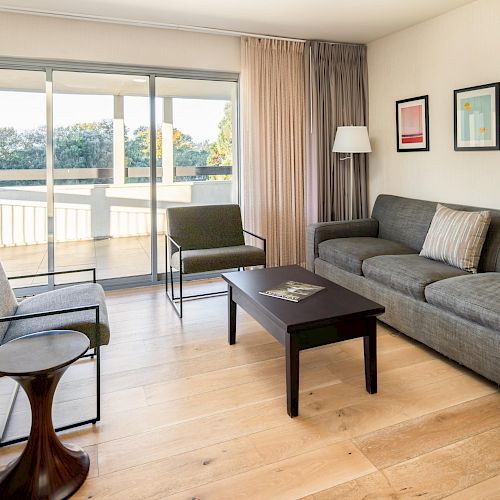 A modern living room with a gray sofa, two armchairs, a coffee table, wall art, and large sliding glass doors leading to a balcony.